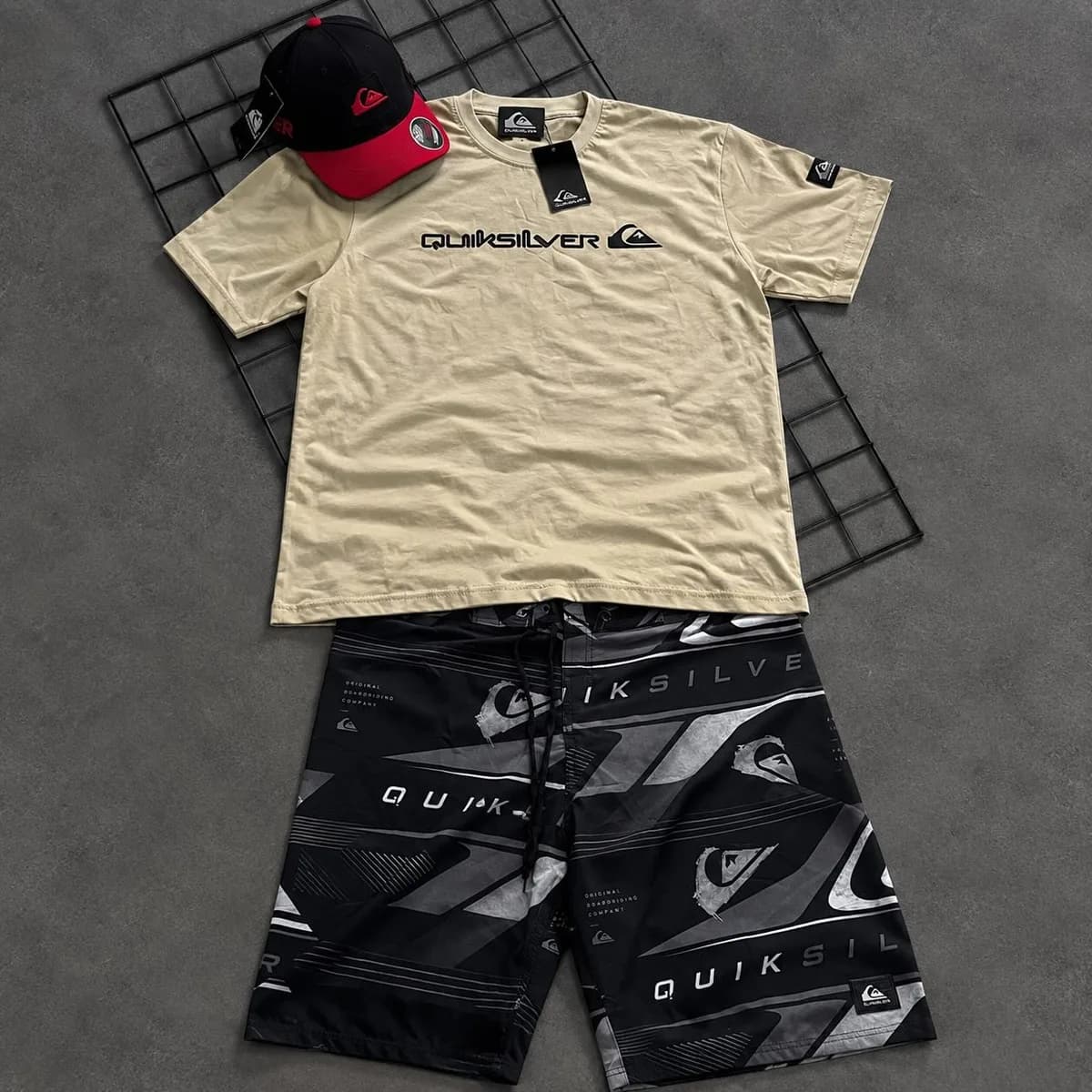 Kit Quiksilver no chão de concreto: camiseta areia, bermuda estampada preta e boné vermelho